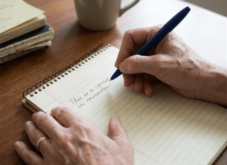 Aparecen las manos de una mujer que escribe sobre una libreta "This is a course in miracles"