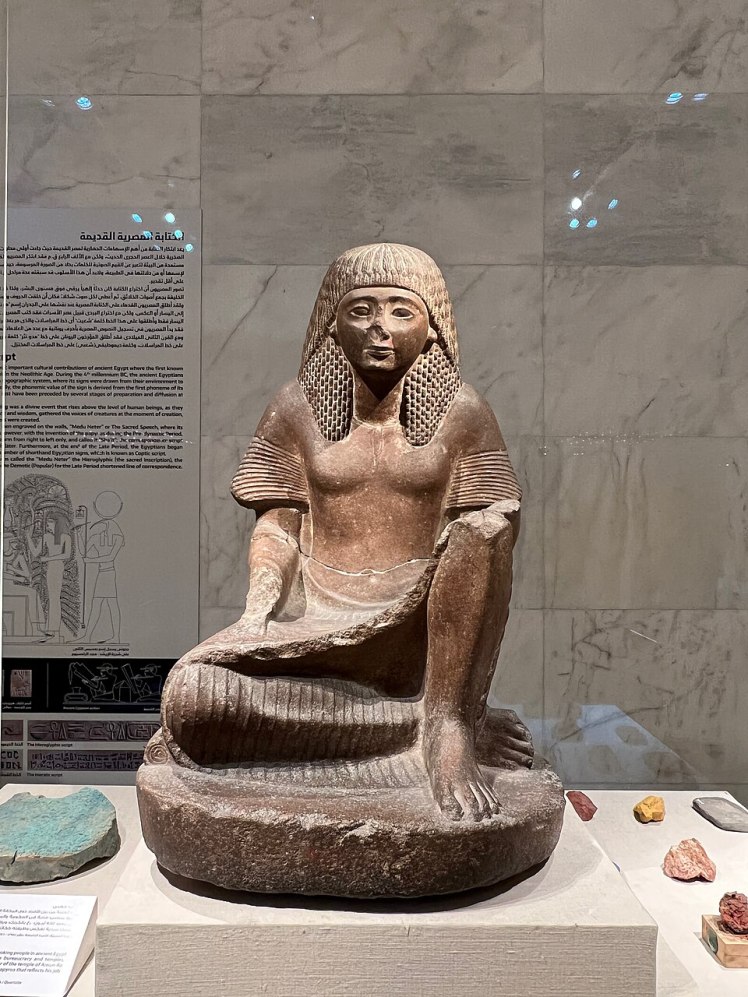 Estatua de Hapi el escriba. Se encuentra con el pecho descubierto, sentado con una rodilla arriba y otra pierna acomodada en el suelo. Tiene un folio de papiro en su regazo.